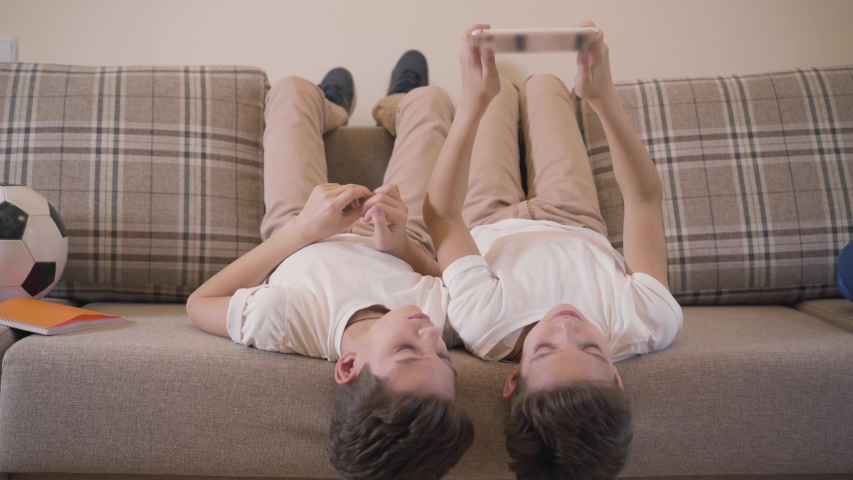 Two cheerful Caucasian twin brothers laying on couch and using tablet. Siblings resting together at home. Children using social networks, game addiction, modern kids.