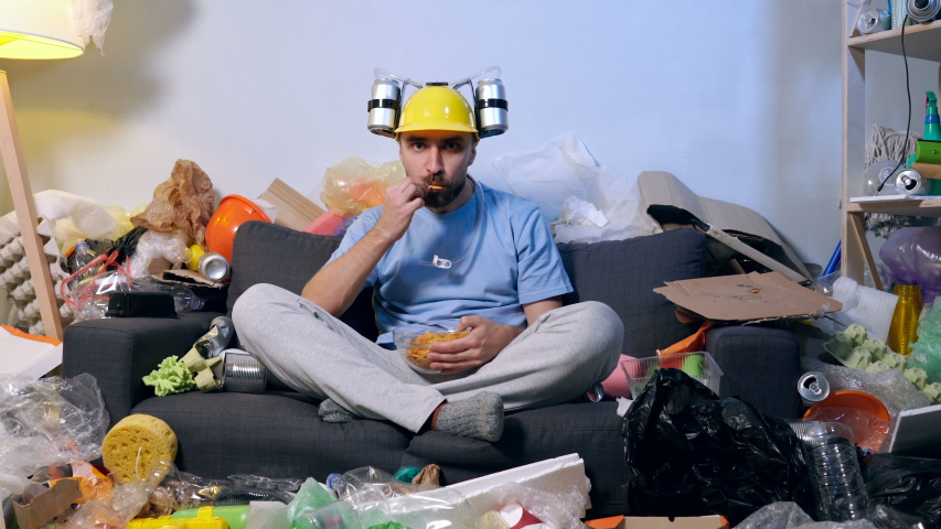 Funny looking man with bear helmet on the had watching football match. Sitting in the room full of trash and garbage. Rejoicing, screaming, victory emotion.
