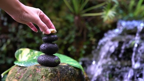 Woman hand put zen stone on a stacked zen stones with garden and mini waterfall background. Zen, spa or wellness concept.