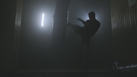 Male boxer training with punching bag on black smoky background with light. Strength and motivation. Silhouette of boxer preparing for big fight. Full length shot in 4K, UHD with copy space