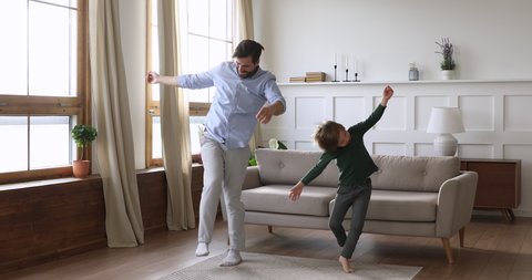 Playful crazy young daddy and cute kid son having fun dancing together in living room interior, happy funny active child boy copy father jumping laughing at home, carefree male family leisure indoors