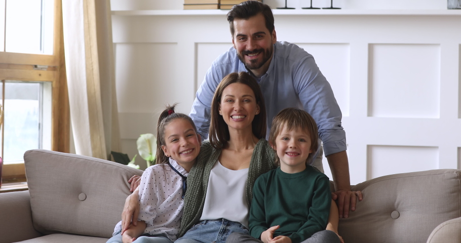 Happy family of four portrait, young adult foster parents mother and father bonding with funny cute school children kids son daughter laughing look at camera posing together on couch in modern home