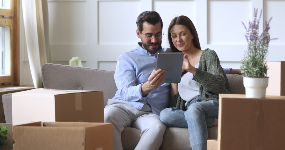 Happy young 30s couple using digital tablet computer sit on sofa on moving day relocate with boxes in new home, man and woman renters choose furniture online removal delivery service in living room Royalty-Free Stock Footage #1041538696