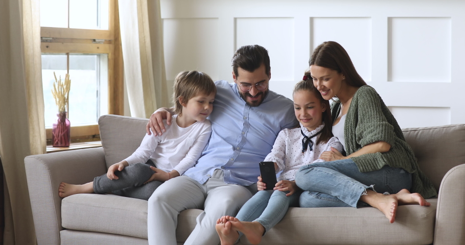 Happy family adult parents with cute school kids children relax on sofa using funny smartphone apps laughing having fun with technology together looking at phone screen take selfie play game at home Royalty-Free Stock Footage #1041538702
