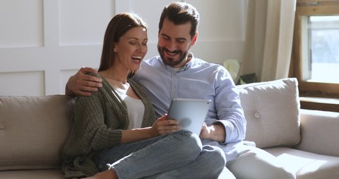 Happy young adult couple bonding laughing using digital tablet sitting on sofa, smiling husband embracing wife holding pad computer surfing internet on couch enjoying family lifestyle at modern home