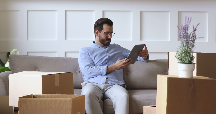 Young man renter or home owner use digital tablet sit on sofa with boxes in new apartment, adult guy choose online removal delivery relocation service interior design renovation idea on moving day Royalty-Free Stock Footage #1041538714