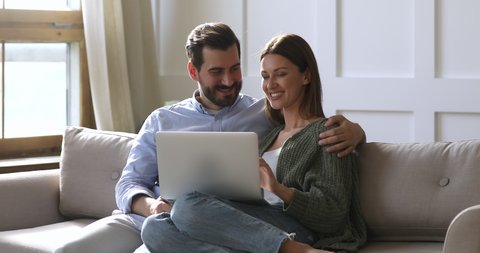 Happy husband and wife relaxing sitting on sofa enjoying using laptop computer, 30s young couple looking at notebook screen browsing internet choosing shopping online resting together in apartment
