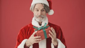 Cheerful elderly gray-haired bearded man in Santa Claus costume is guessing what inside the box isolated over the red background in studio