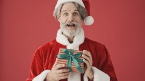 Happy elderly gray-haired bearded man in Santa Claus costume is presenting a gift to the camera isolated over the red background in studio