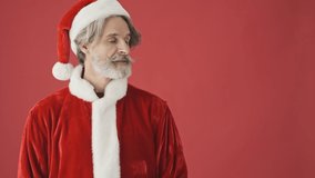 Attractive gray-haired bearded man in Santa Claus costume looking to the side and showing something with his empty hands isolated over the red background in studio