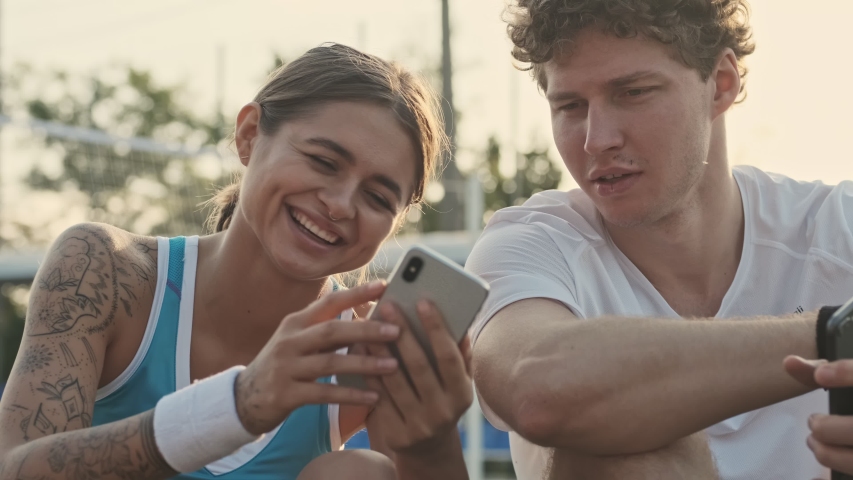 Happy curly male tennis player having rest and using smartphone with his smiling female opponent on the sports ground | Shutterstock HD Video #1041540400