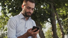 Happy bearded businessman answering the call and talking by smartphone while sitting in park outdoors