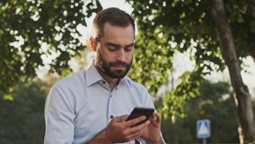 Calm bearded businessman using smartphone while sitting in park outdoors