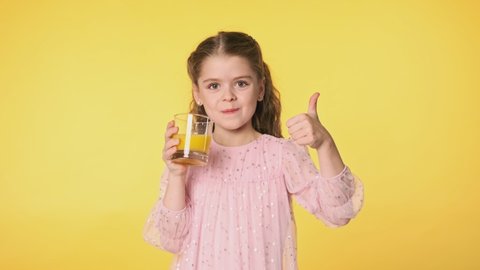 Medium portrait of child with glass of juice. Beautiful kid girl drinking orange juice from glass at yellow background. Slow motion. 4K, UHD