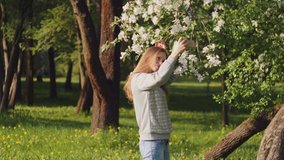 Pretty happy girl is making a selfie with a smartphone on background of a blooming apple tree at the spring park. Concept of woman portrait photography, gadgets or travelling. 4K slow motion video.