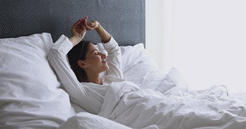 Happy healthy young adult woman waking up on early pleasant lazy weekend lying in cozy bed on soft pillow orthopedic mattress stretching raising arms enjoy good morning concept feel fresh and rested