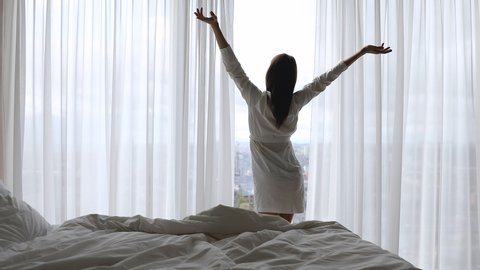 Happy young woman wearing dressing gown get up from bed come to big window open curtain lace look outside at beautiful view in modern hotel bedroom in morning apartment enjoy luxury lifestyle concept