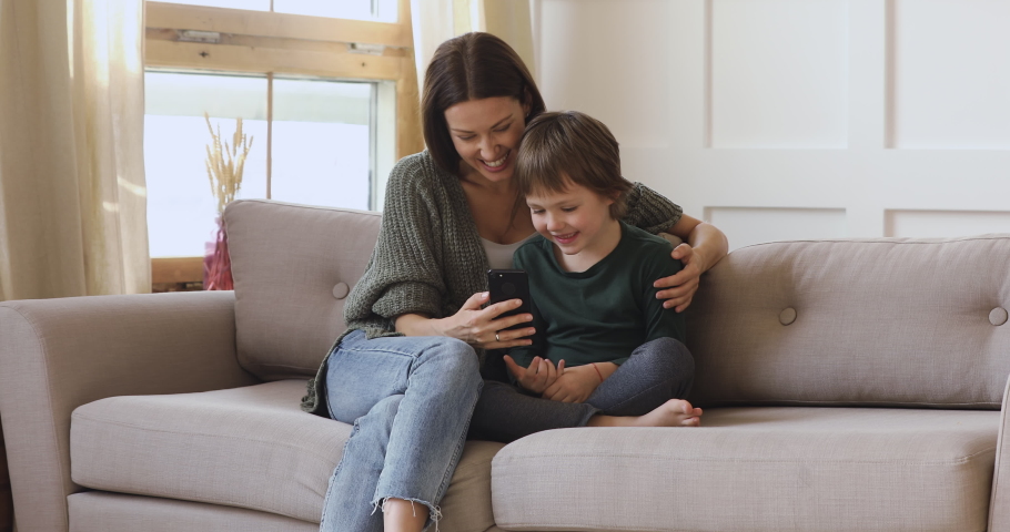 Happy young adult mum teaching cute kid son learning using smartphone funny apps at home, smiling mother hug preschool child boy having fun looking at cellphone relax with technology gadget on sofa Royalty-Free Stock Footage #1041547624
