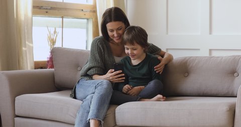 Happy young adult mum teaching cute kid son learning using smartphone funny apps at home, smiling mother hug preschool child boy having fun looking at cellphone relax with technology gadget on sofa