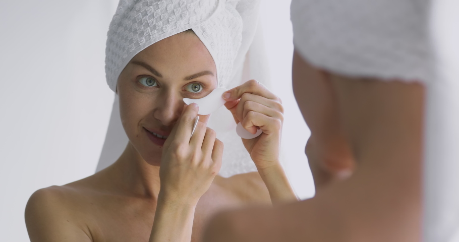 Happy attractive 30s woman apply eye care patches looking in bathroom mirror, healthy beautiful adult lady put undereye face skin care moisturizing pads for dark circles bags spa treatment concept Royalty-Free Stock Footage #1041547762