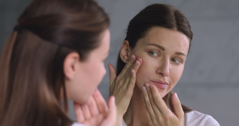 Worried young adult 30s woman looking in mirror feeling stressed about facial wrinkles problem, frustrated upset lady touching face depressed about dry sensitive aging skin care concept in bathroom | Shutterstock HD Video #1041547765