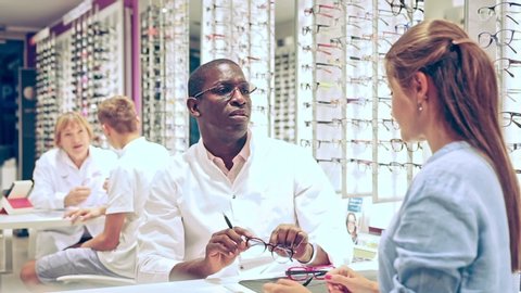 African-American optometrist consulting young woman in optical store, helping to choose eyeglasses