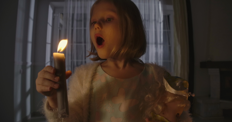 Close-up portrait of strange Caucasian girl blowing out candle and holding doll in other hand. Weird child alone at night. Scare, fear, strangeness.