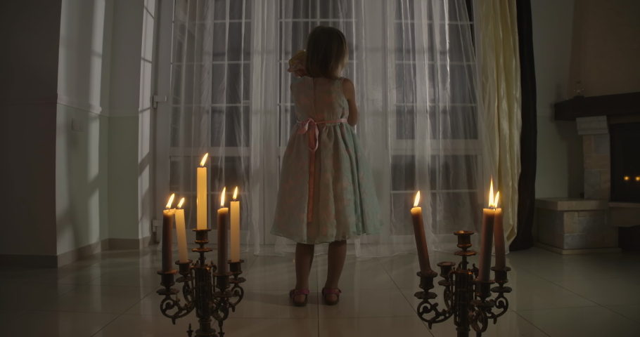 Camera approaching little Caucasian girl standing in candlelight and holding doll. Weird child caressing her toy and looking around. Scare, fear, strangeness.
