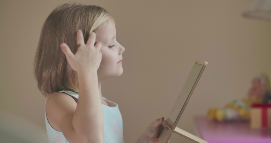 Side view of blond Caucasian girl looking at the mirror, smiling and fixing her hair. Funny child in blue dress with pink butteflies making faces.