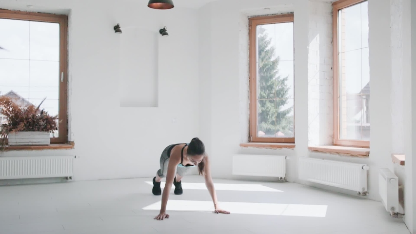 Cameraman shooting fit young brunette girl in sportswear running plank on floor. Videographer shoots sporty female doing exercizes in bright studio, backstage of making video about healthy lifestyle