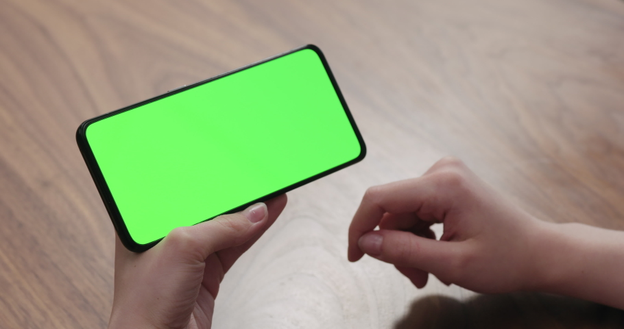 Young woman sitting at a table and using a smartphone with horizontal green screen