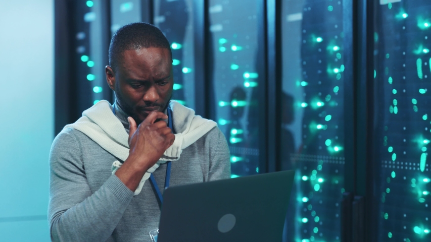 Pensive black IT specialist working on laptop shaking head thinking solving a problem. Satisfied server technician finding a valuable solution smiling coworking in data center server room. | Shutterstock HD Video #1041568234