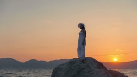 A woman in a white dress standing on a large rock in the water on a background of the sun and wind. RAW video record.