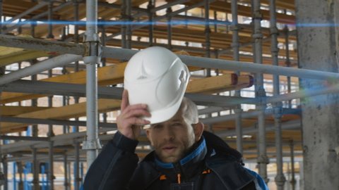 Caucasian worker or builder gets ready for work and puts on white helmet while walking on construction site. Safety precautions, starting workflow, anamorphic shot, lens flare