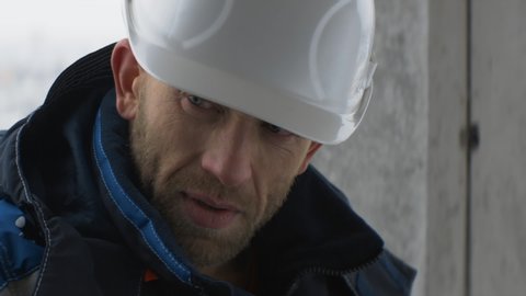Tired caucasian worker or foreman in blue uniform takes off white helmet and wipes sweat from his forehead inside an unfinished building. Fatigue, feeling unwell, sadness, anamorphic shot