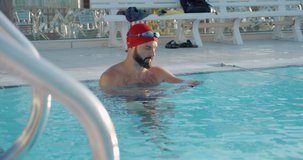 Muscular young man in red cap in swimming pool starting his training with heart rate monitor at the sunny day. Slow motion 4k video