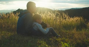 Happy traveler father and son sitting sitting on grass and look through binoculars, bonding around mountain under sun light enjoying the leisure and freedom. 4K slow motion video