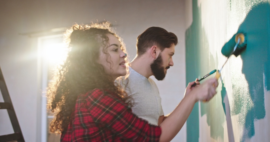 Cute and beautiful young Caucasian woman painting walls in the apartment in blue color with a wall brush roller and having fun with her husband, laughing and painting on each other's faces. Royalty-Free Stock Footage #1041592264