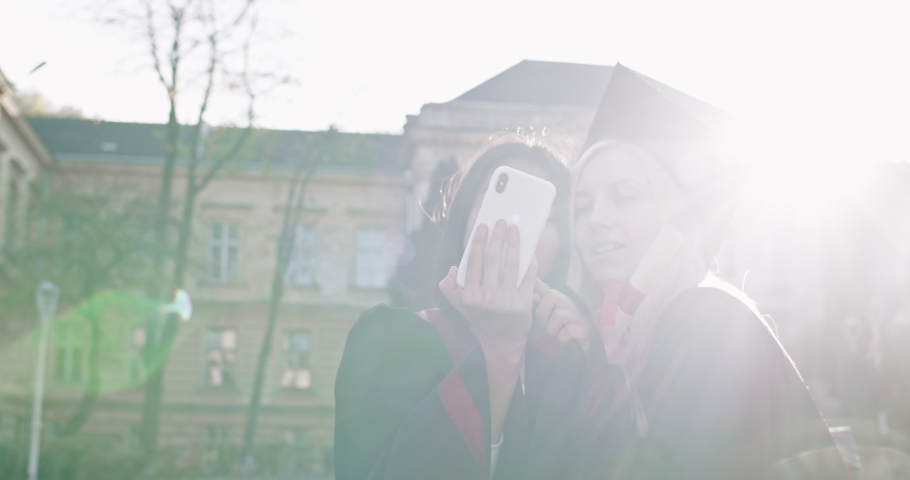 Close up of the two multiethnic young best friends girls, pretty master graduates with diplomas and in academic caps. One girl showing to another photos, chat or video on the smartphone. Outdoors.