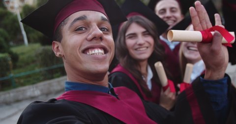 Close up of the moulatto young joyful guy in academic cap and gowns taking funny selfie photos with his funny and hppy friends students at their graduation day. POV. Outside.