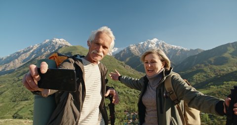 Mature caucasian couple travelling together, having a nordic walking hike in spring mountains, then stopping to take a picture on smartphone, spending time after retirement - recreational pursuit