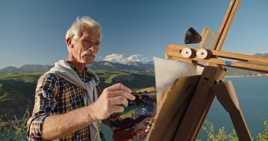 Old caucasian man standing in front of easel, drawing a picture, inspired by beautiful view of lake in mountains. Senior man enjoying his hobby after retirement - pension, recreational pursuit 4k