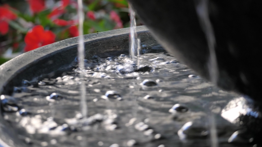 Close up slow motion panning clip of water trickling down into a pool creating bubbles and splashing in a black tiered granite round or bowl shaped fountain with red flowers blurred in the background.