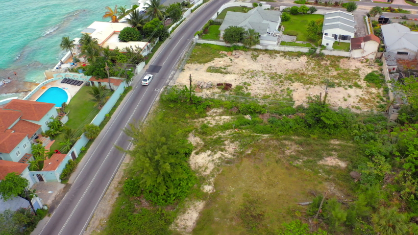 Aerial: White Car Driving Along Waterfront Road Past Tropical Beach Homes - Nassau, Bahamas