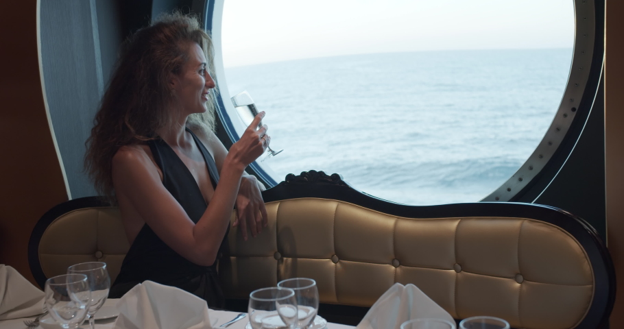 Beatiful woman is sitting alone at table in restaurant on cruise ship, drinking champagne.