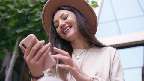 Pretty cheerful 30-aged lady with straight brown hair in beige fashion hat watching funny photos on her mobile and wholeheartedly smiling Adlı Stok Video