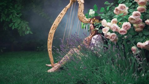 Fantasy Woman. white vintage short dress, bare legs. Pretty Goddess sits in garden with old harp.  blooming roses hydrangeas. gold laurel wreath. princess enjoying nature. musician fashion model