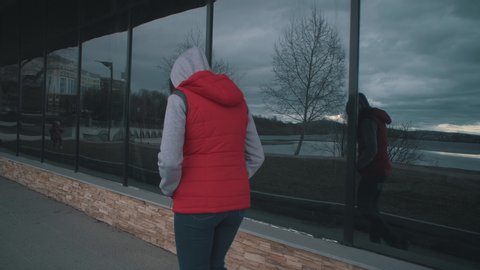 View from the back of young brunette woman in red sleeveless jacket is walking alongside large windows on cloudy day.Stooped, lost girl is reflected in glass of building, tracking shot.