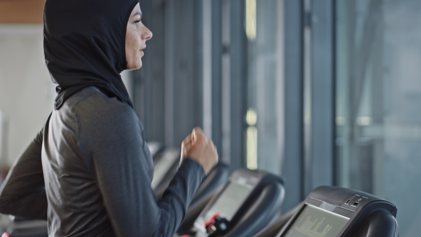 Athletic Muslim Sports Woman Wearing Hijab and Sportswear Running on a Treadmill. Energetic Fit Female Athlete Training in the Gym Alone. Urban Business District Window View. Back View Arc Shot Royalty-Free Stock Footage #1041637402