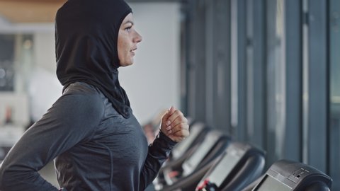 Athletic Muslim Sports Woman Wearing Hijab and Sportswear Running on a Treadmill. Energetic Fit Female Athlete Training in the Gym Alone. Urban Business District Window View. Back View Arc Shot